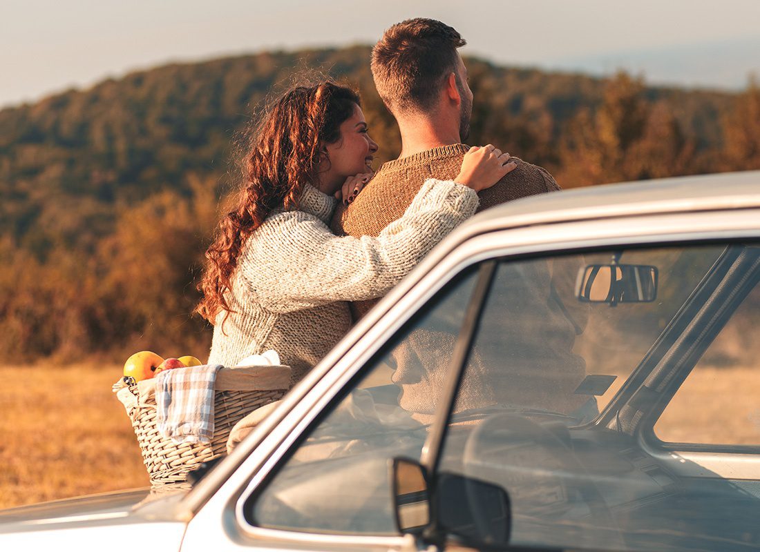 About Our Agency - Rear View of a Young Couple Sitting on the Front of an Old Car with a Picnic Basket Enjoying the Views of the Mountains