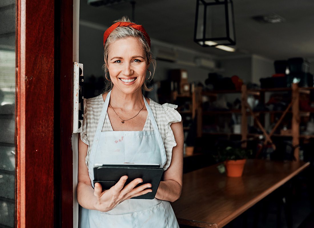 Business Insurance - Closeup Portrait of a Smiling Mature Woman Standing Outside the Front Door of her Small Main Street Shop While Holding a Tablet